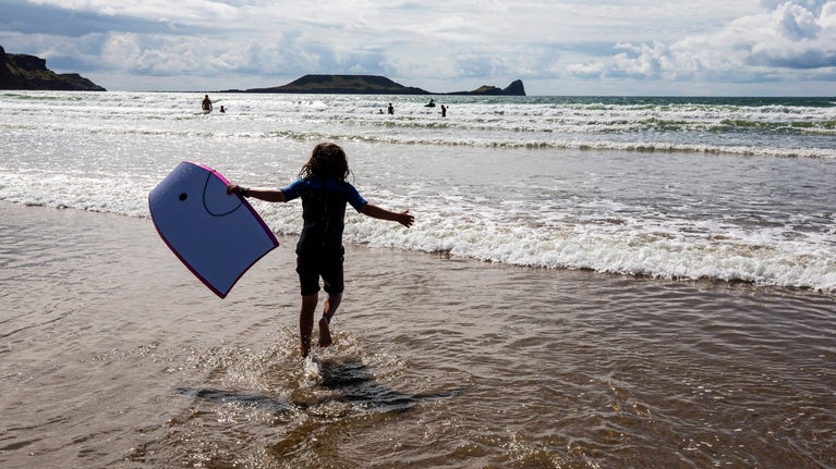 The silhouette of a child running into the sea with a bodyboard at Rhosili and South Gower Coast, Wales